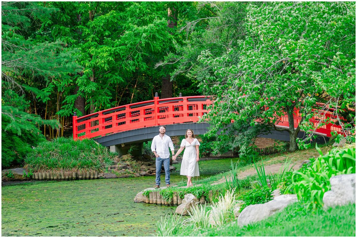 A loving couple holding hands next to a beautiful waterway and iconic red bridge in their Duke Gardens engagement photos by JoLynn Photography