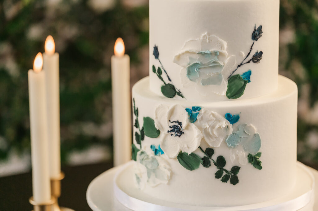 A white wedding cake with blue flowers and warm lighting at The Atrium Wedding Venue by JoLynn Photography