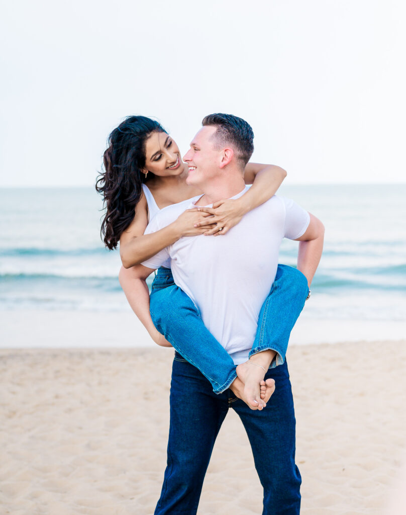 A couple playing piggy back riding on Wrightsville beach for their Wilmington engagement photography session by JoLynn Photography