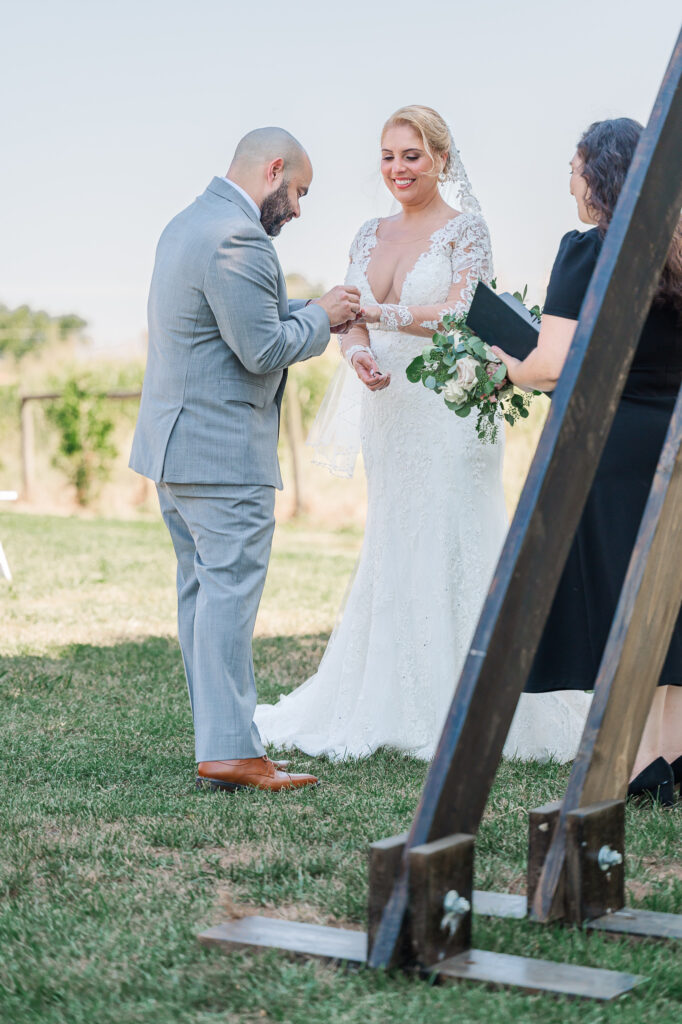 A groom placing a wedding band on his wife's hand at The Boots and Roots Farm by JoLynn Photography