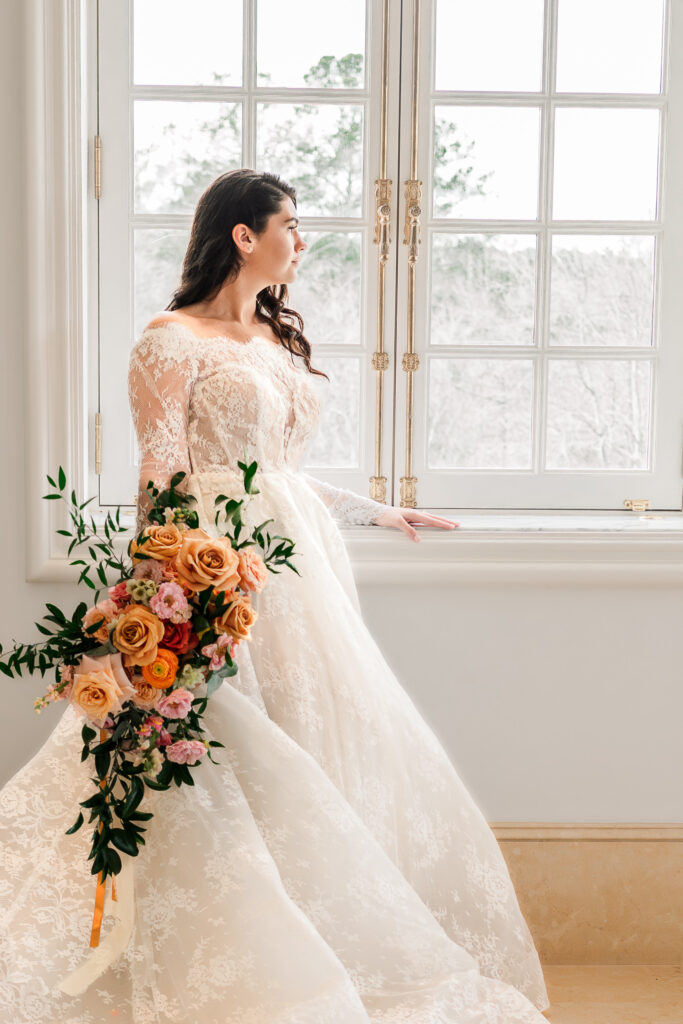 A brunette bride looking out a window at the Oxbow Estate by JoLynn Photography