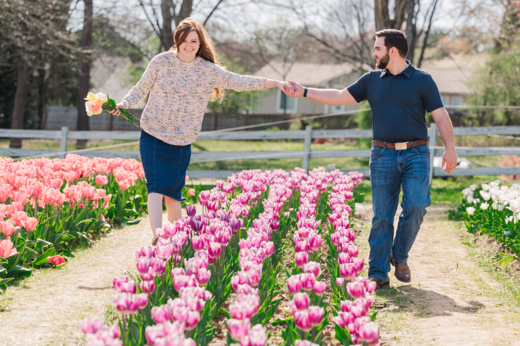 An engaged couple walking and holding hands over tulip fields by JoLynn Photography, a Greensboro wedding photographer
