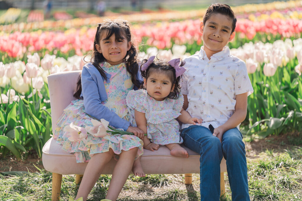 Three happy children sitting on a bench in a field of tulips by JoLynn Photography