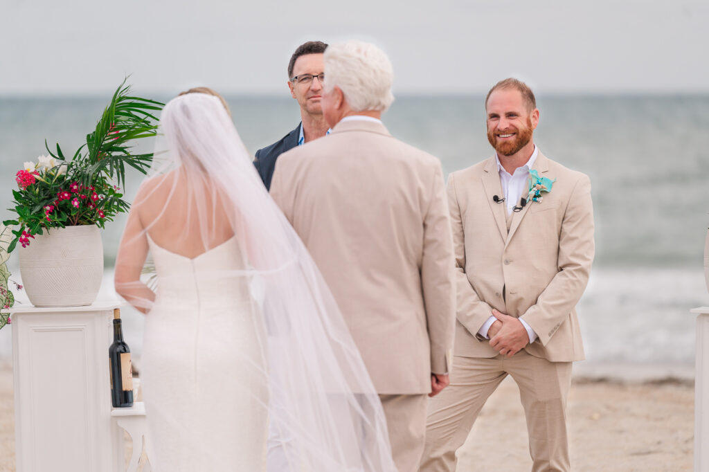 A groom smiling at his soon-to-be wife with her dad as he gives her away on her Atlantic beach wedding day by JoLynn Photography