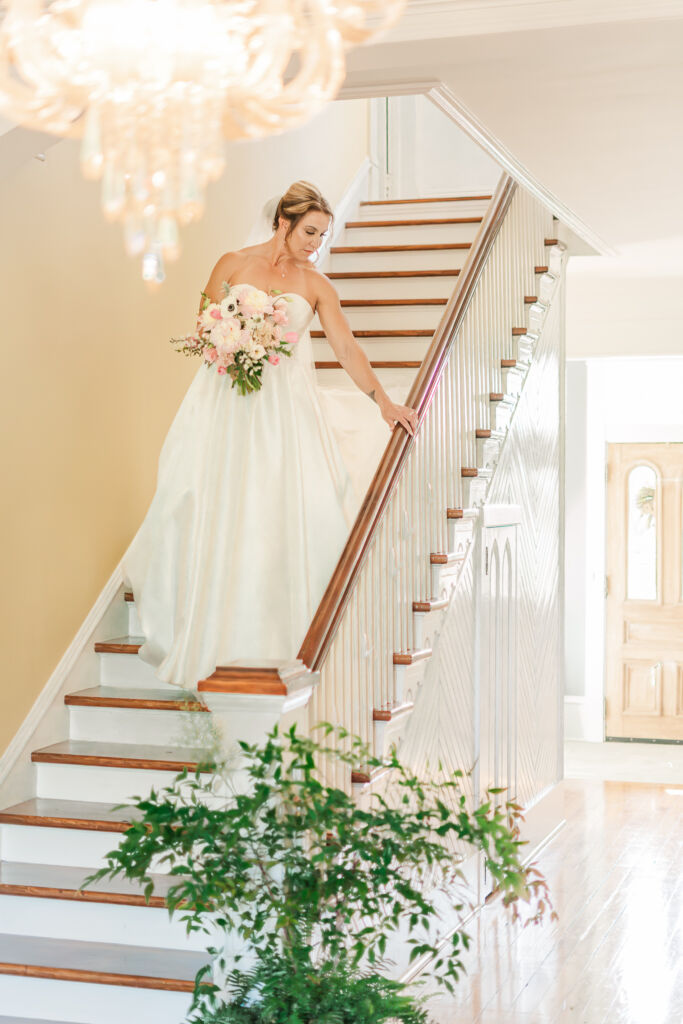 A happy bride walking down a grand staircase with a chandler at Highrock Farms by JoLynn Photography