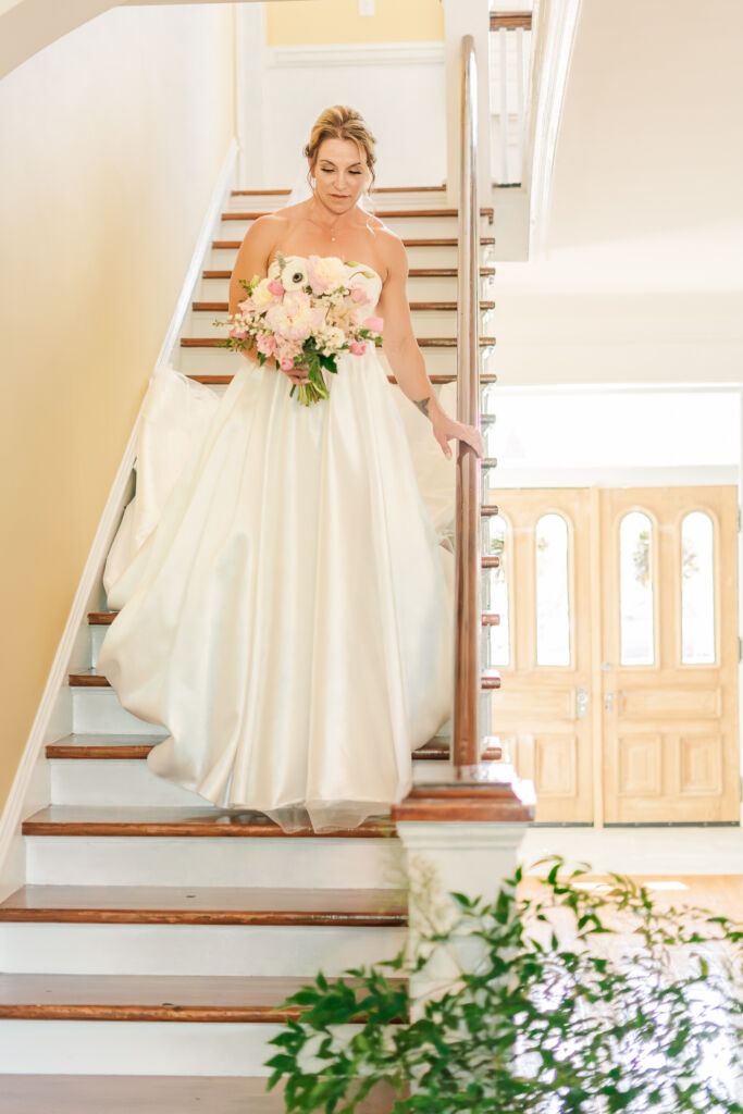A happy bride walking down a staircase at her wedding venue, Highrock Farms, holding her summer wedding bouquet during her bridal portrait session