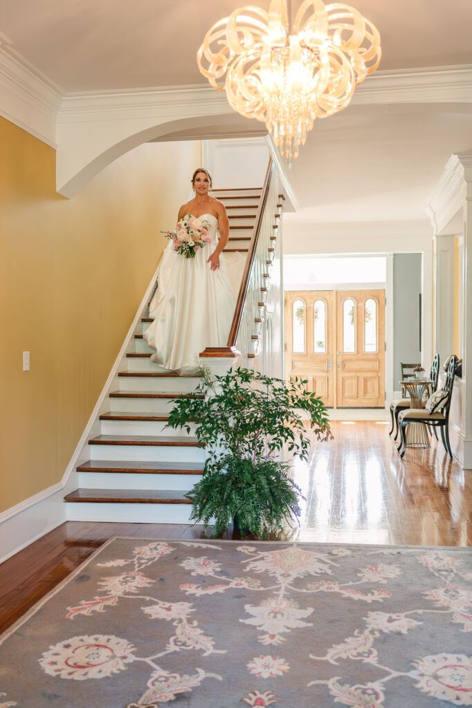 A smiling bride walking down a grand staircase with a bright chandler at Highrock Farms for her bridal portrait session with JoLynn Photography