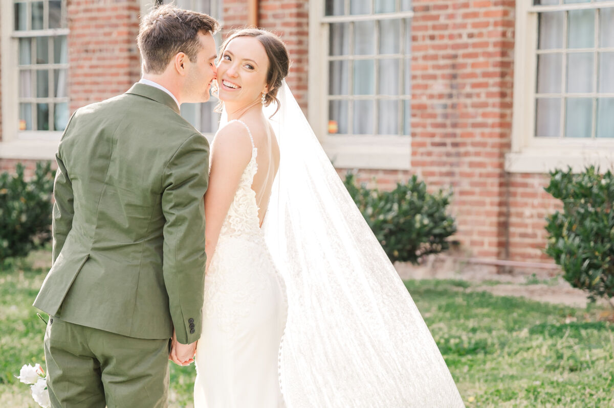 A loving couple holding hands outside of the Woolen Mill Fredericksburg during sunset next to a row of green bushes and warm orange bricks by JoLynn Photography