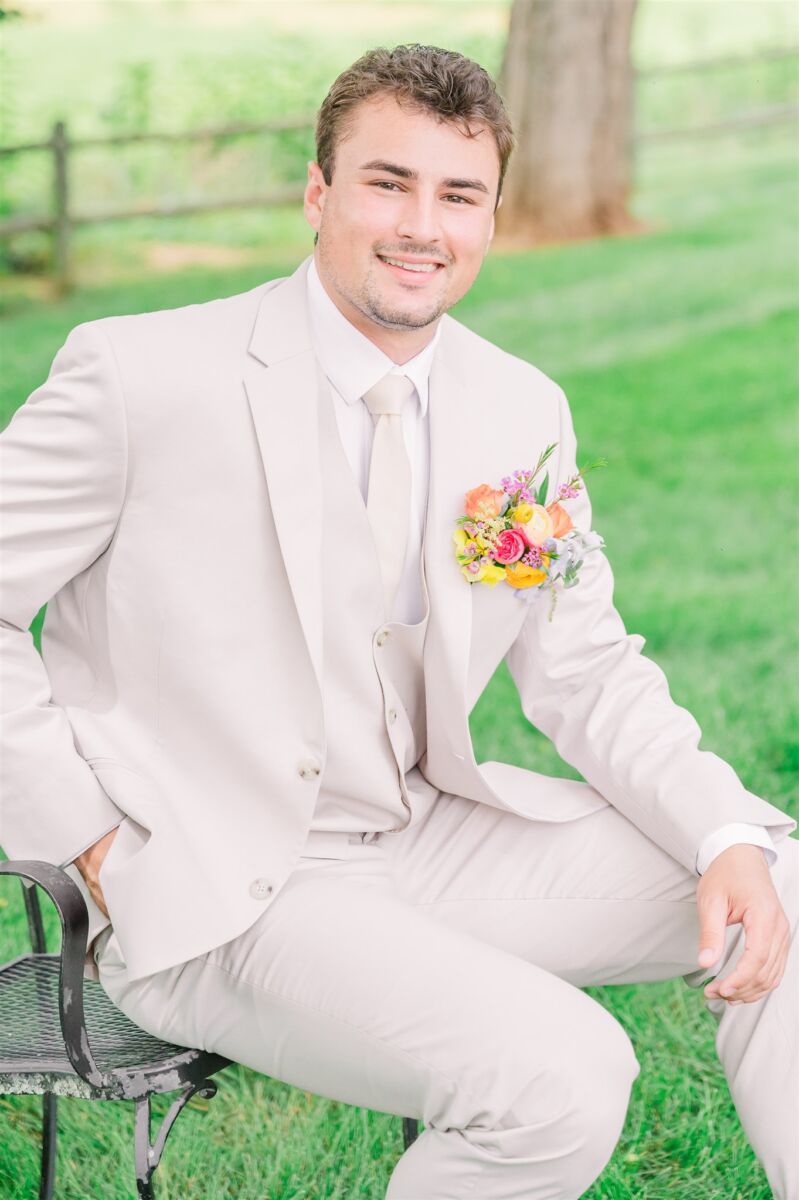 A smiling groom sits on a patio chair in his tan suit in a lawn with a colorful boutonniere