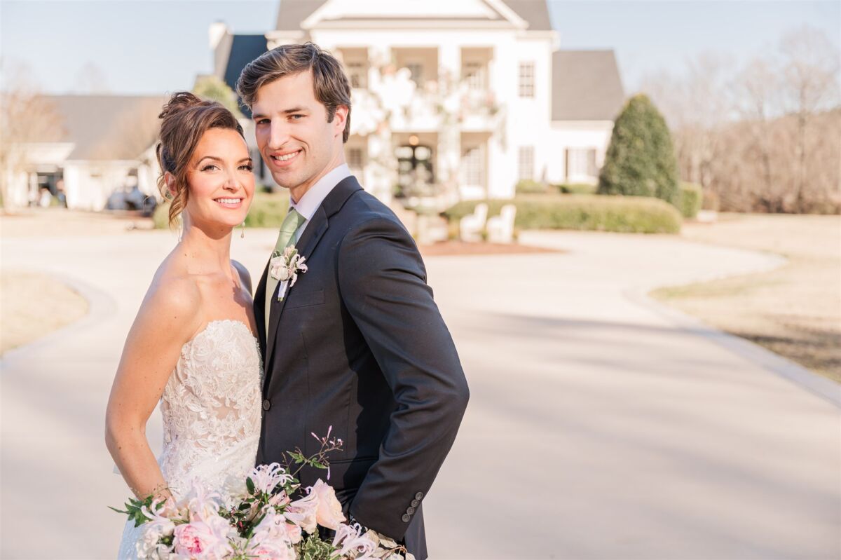 A bride and groom stand chest to chest smiling in a large driveway in front of an estate