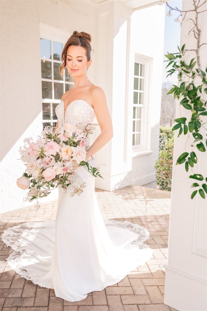 A bride smiles down to her pink bouquet in her hand while standing on a front porch