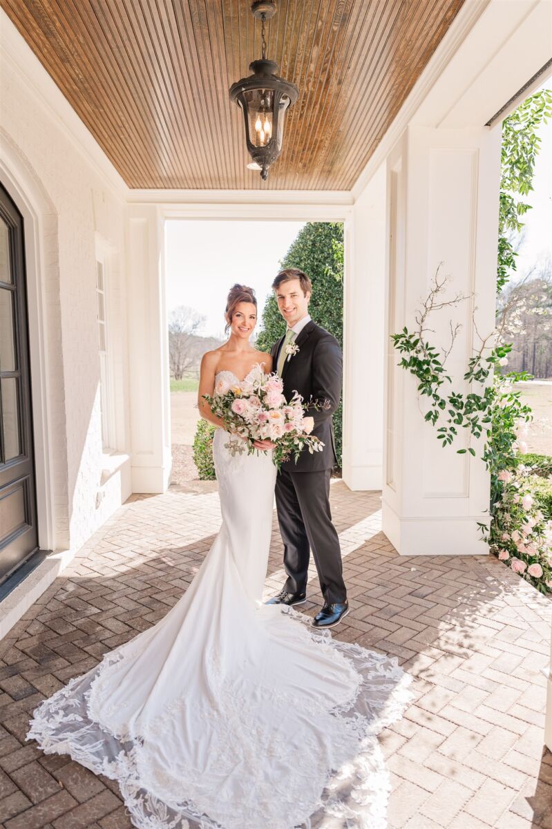 A bride and groom stand behind the long train on a front porch while smiling and holding a large pink bouquet during their Highgrove Estate wedding