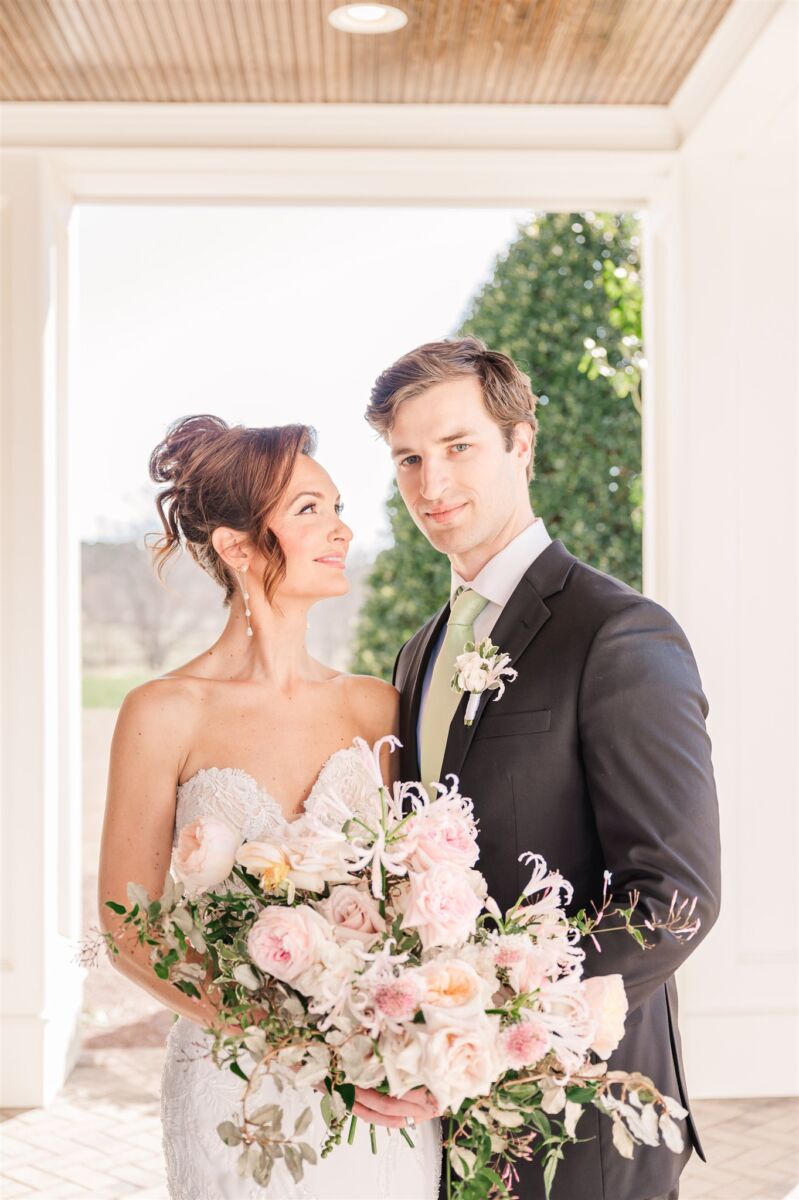 Newlyweds stand smiling at each other in a black suit and lace gown on a porch at their Highgrove Estate wedding