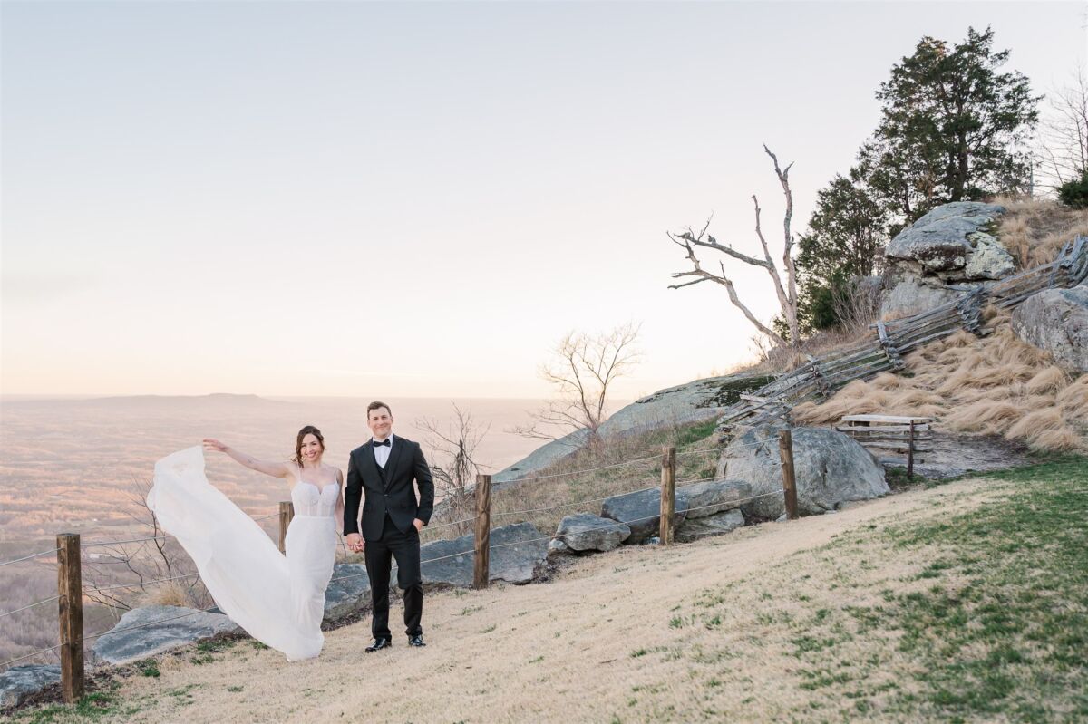 A bride and groom smile on the top of a mountain at sunset holding hands and the train blowing in the wind