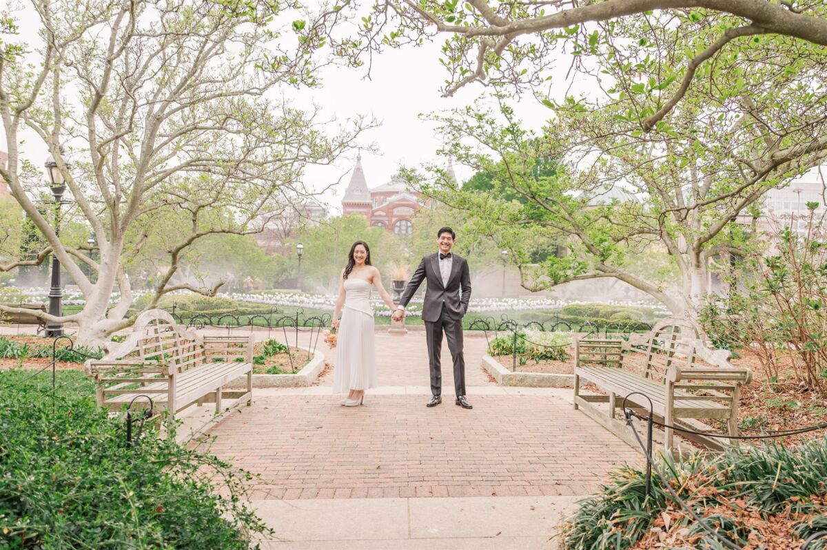 A bride and groom hold hands while walking through a brick paved garden