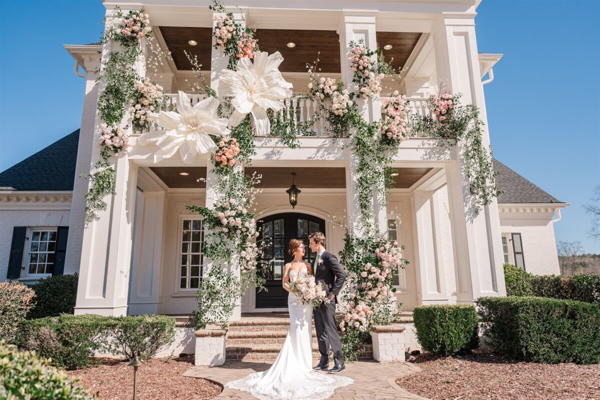 Newlyweds stand under a flower covered front entrance to a white wedding venue smiling at each other thanks to Raleigh wedding planner
