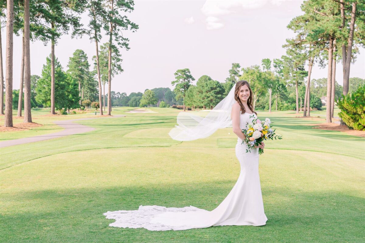 A smiling bride stands on a golf tee as her veil flows in the wind behind her thanks to support from a Raleigh wedding planner