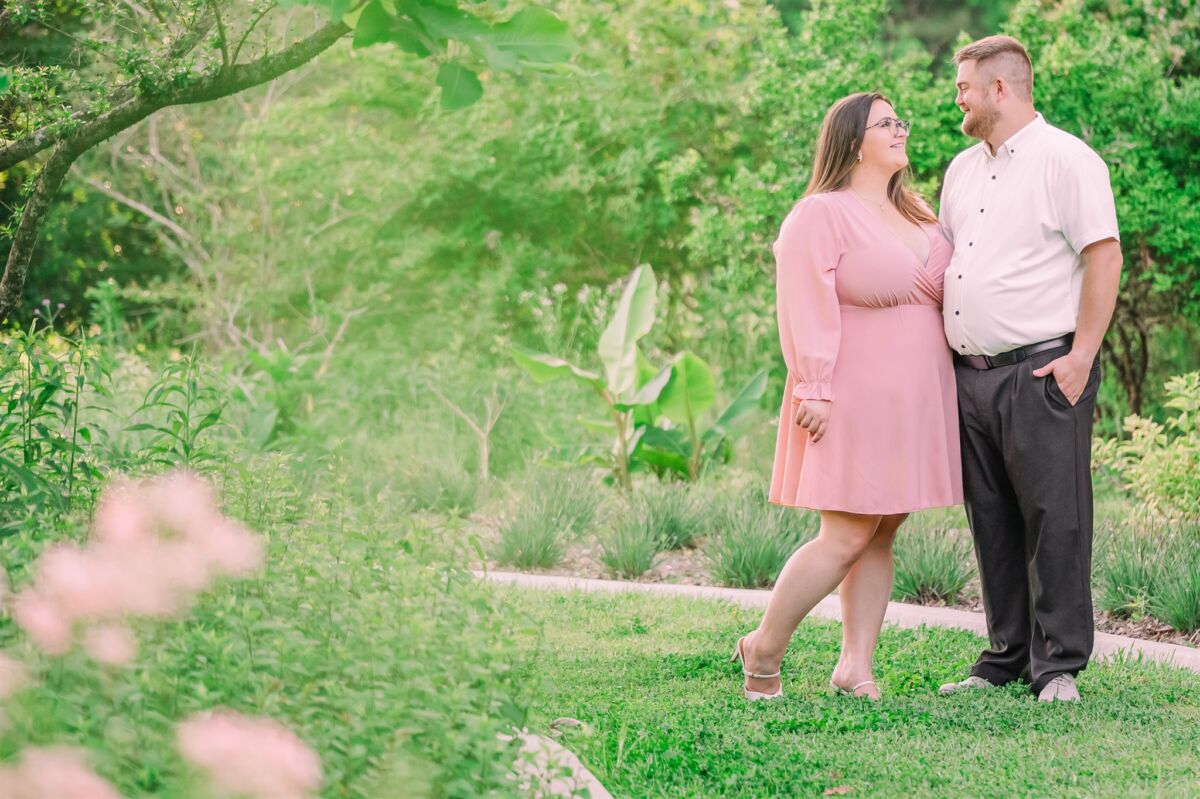A happy engaged couple snuggle and smile at each other in pink and black in a garden