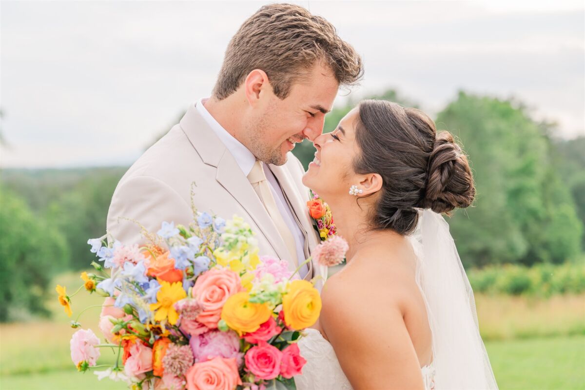 A bride and groom nuzzle noses with big smiles in a meadow with a bright vibrant bouquet