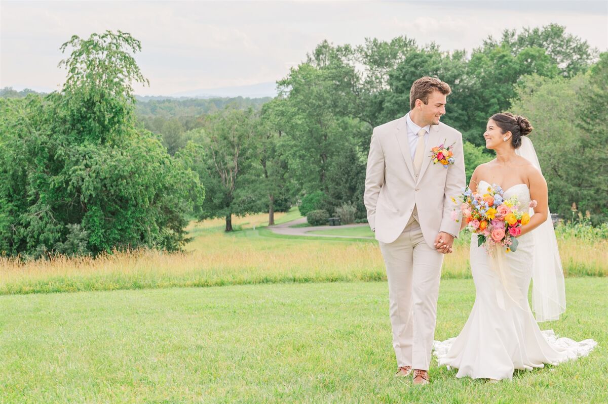 Newlyweds walk holding hands and smiling at each other in a meadow at one of the Apex wedding venues