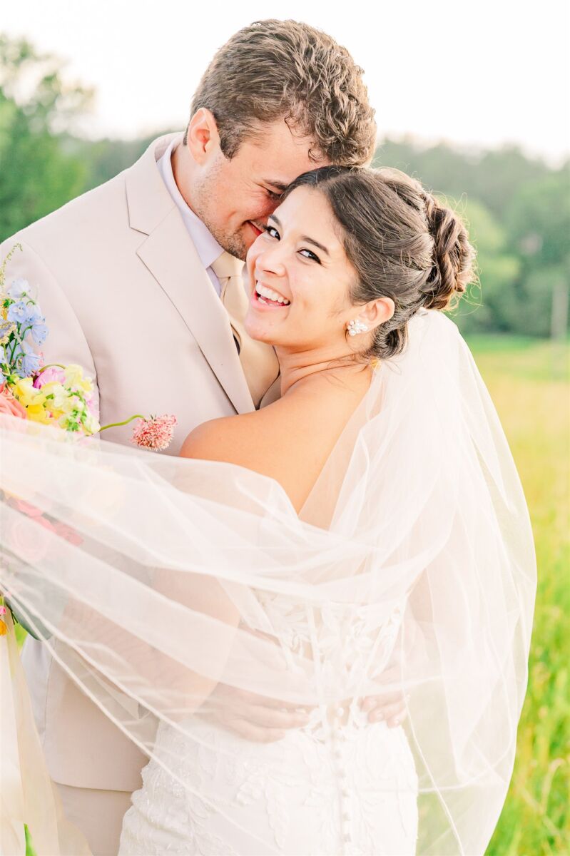 A happy bride smiles over her shoulder while dancing with her groom in a tan suit in a meadow at sunset at one of the Apex wedding venues