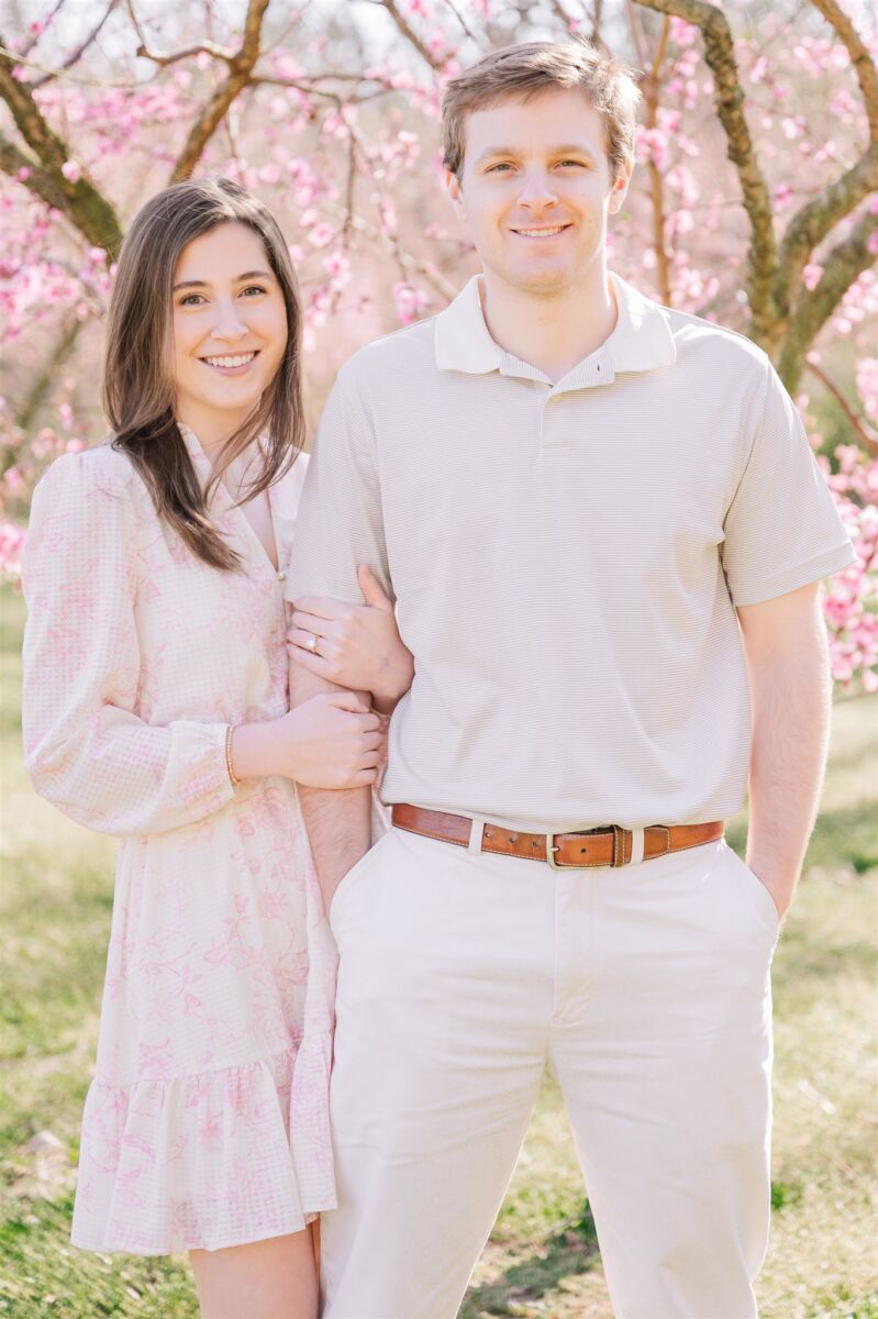 A happy woman in a pink dress holds onto the arm of her fiancee in white in cherry blossoms after finding Greensboro wedding planners