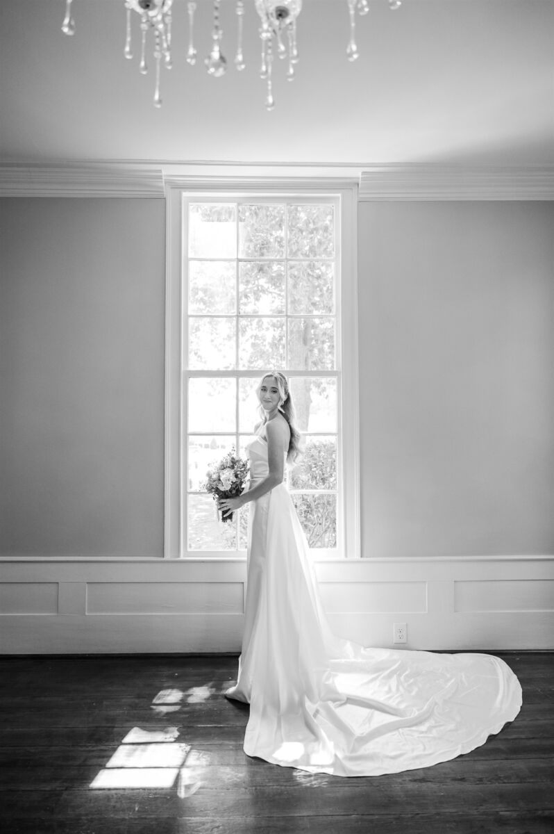 A black and white image of a bride smiling over her shoulder while standing in her silk gown in a window
