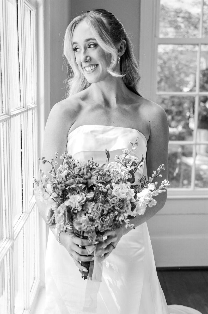 A happy bride smiles out a window while holding her bouquet in black and white at one of the Holly Springs wedding venues