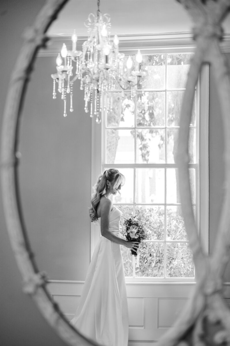 A peek at a bride through a mirror as she stands in a window smiling down to her bouquet at one of the Holly Springs wedding venues