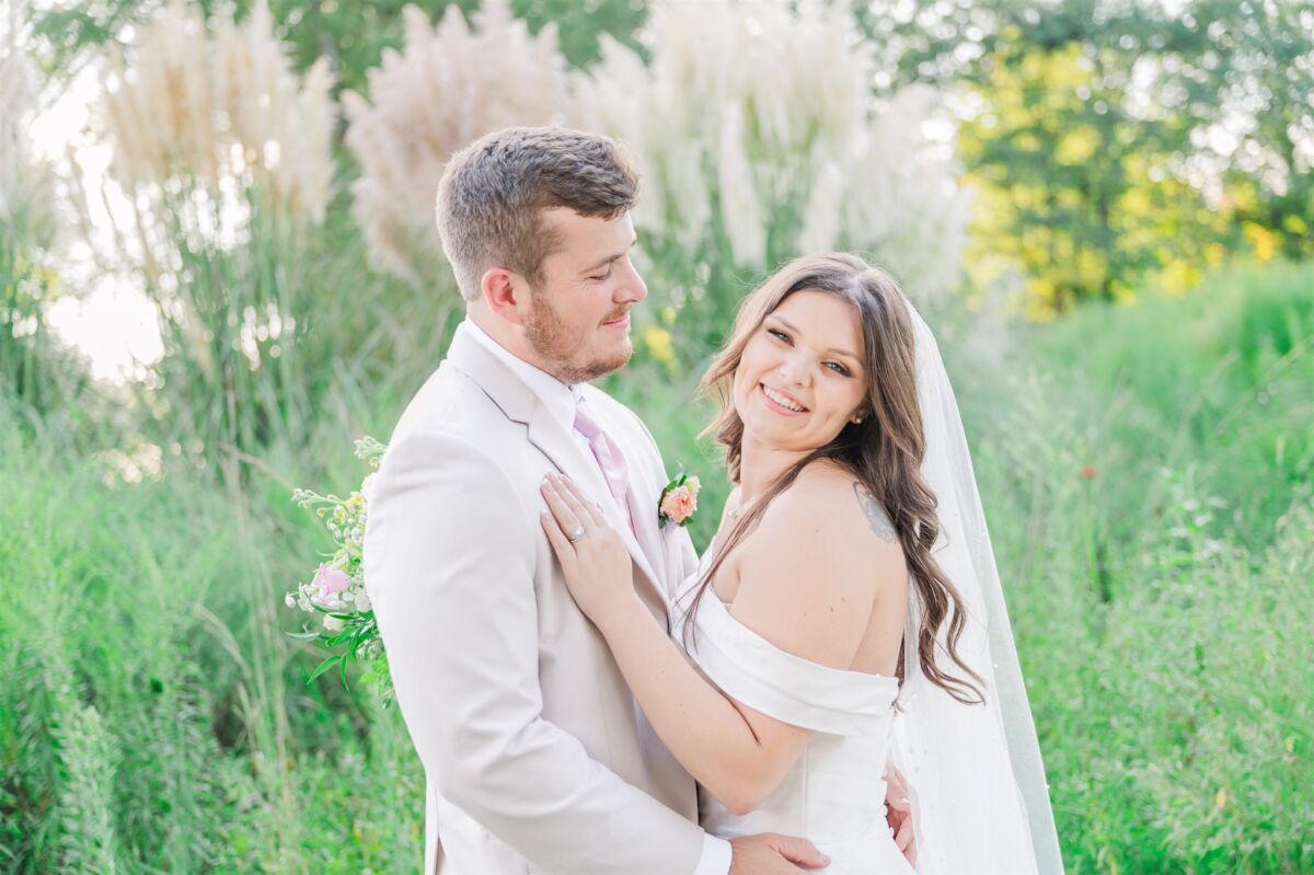 A happy bride smiles over her shoulder while cuddling her groom in a meadow at sunset with pompous grass at one of the Raleigh outdoor wedding venues
