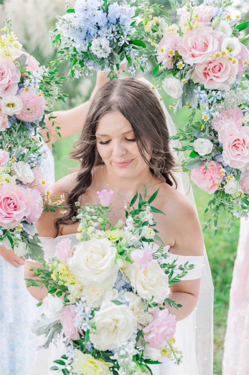 A smiling bride looks down at her bouquet as her bridesmaids hold their bouquets around her head at one of the Raleigh outdoor wedding venues
