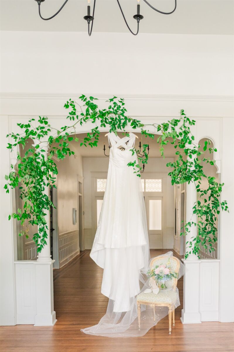 A gown hangs from an archway in a hall with the veil over the bouquet on a chair