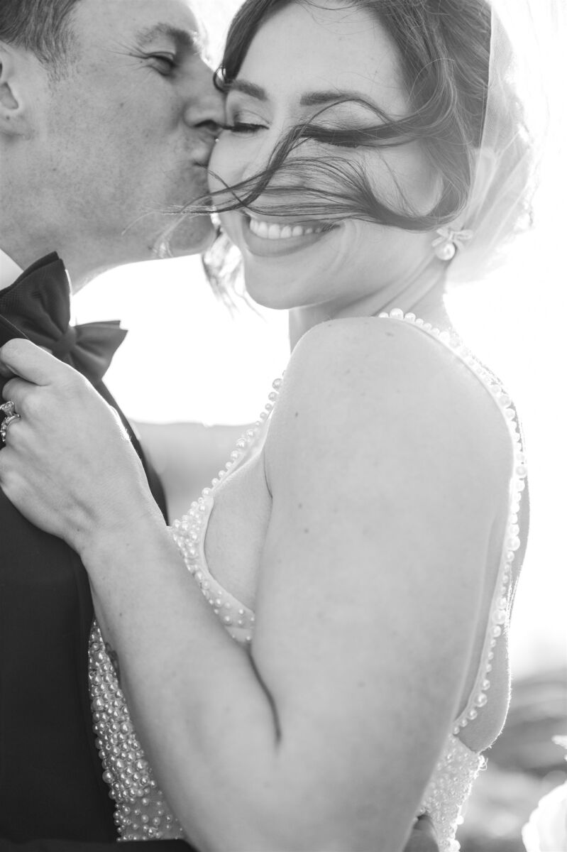 A happy bride is kissed on the cheek in her pearl crusted gown at sunset in black and white at The Club At 12 Oaks wedding venue