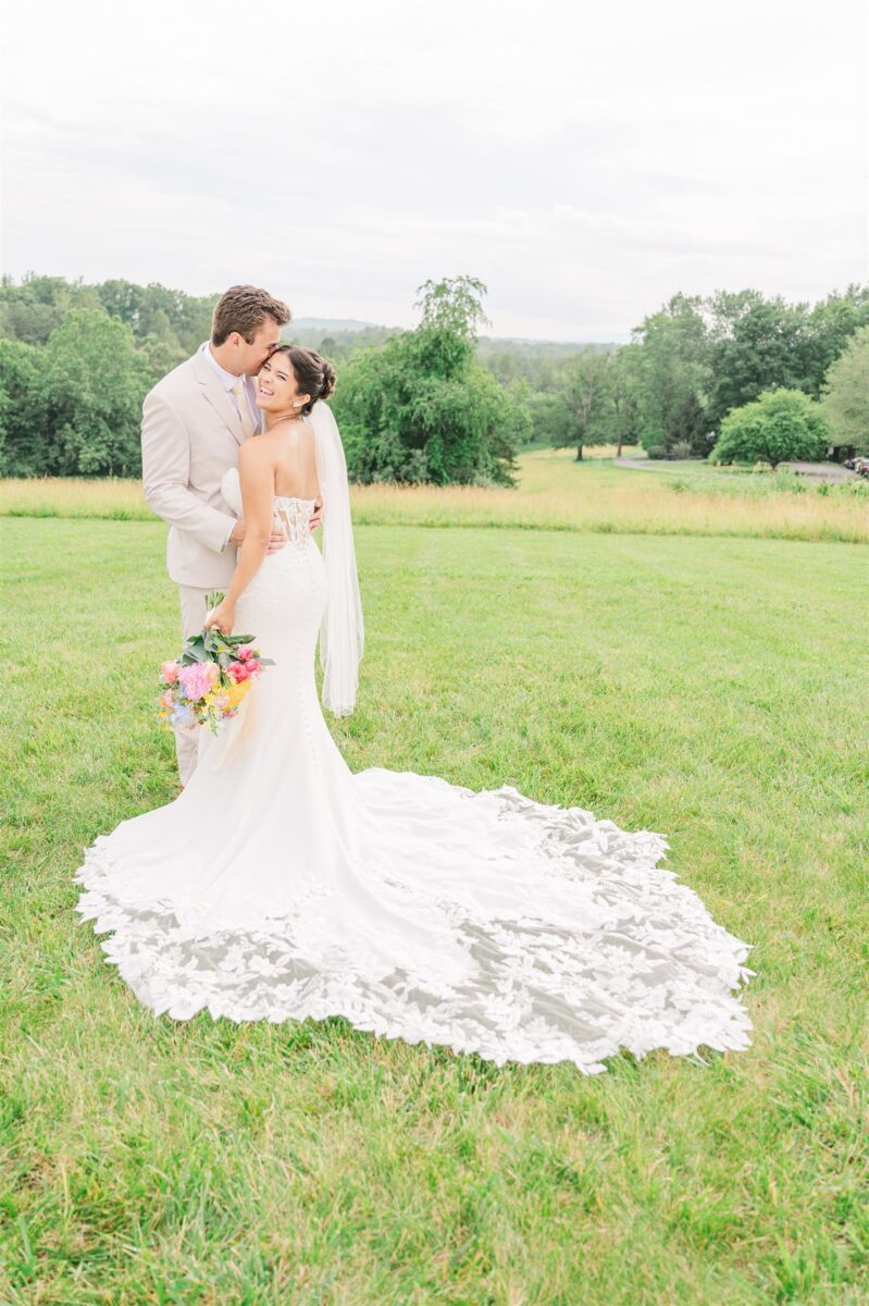 A laughing bride looks over her shoulder while cuddling her groom in a tan suit in a meadow at The Meadows At Firefly Farm Preserve
