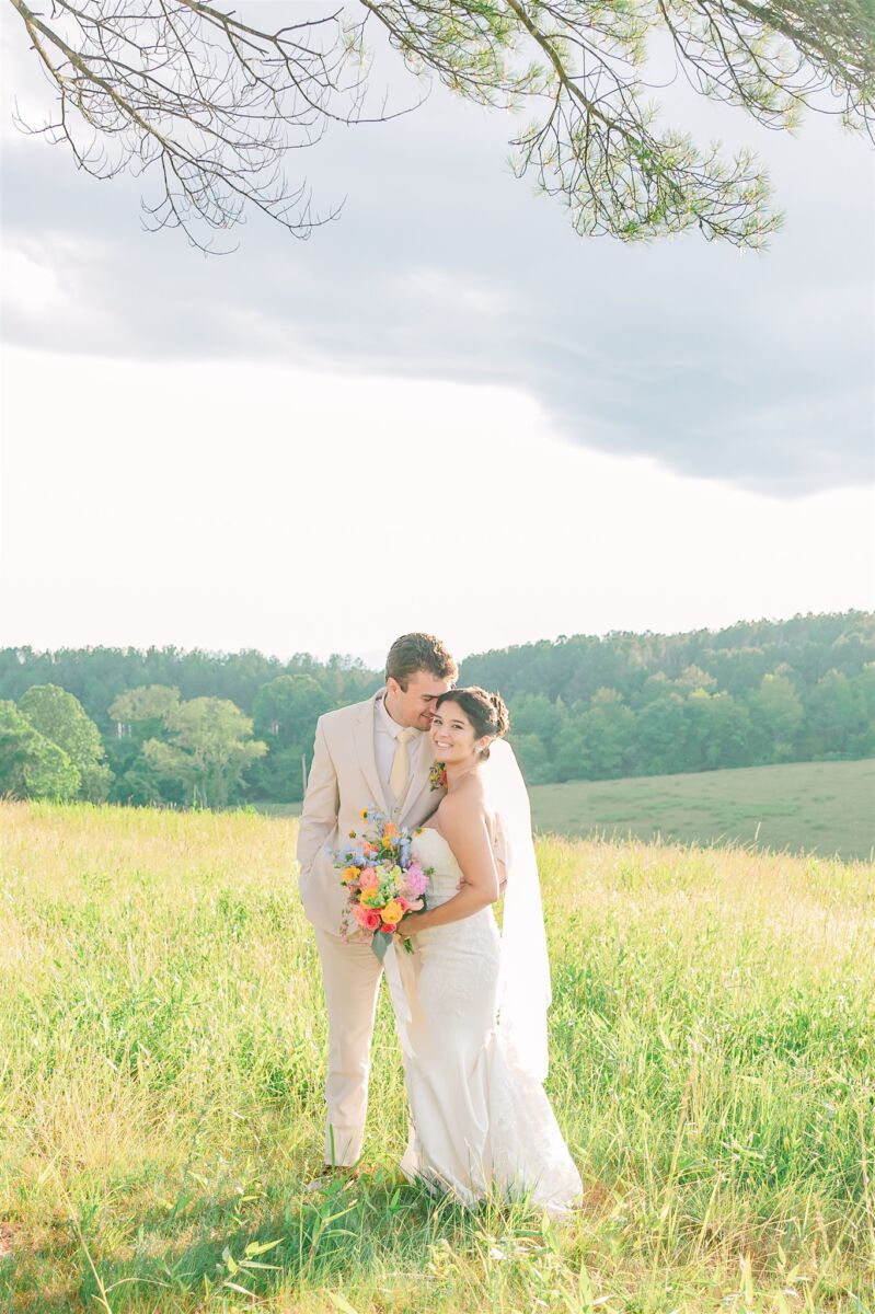 Newlyweds snuggle and smile in a grassy hill in a cream suit with a large colorful bouquet