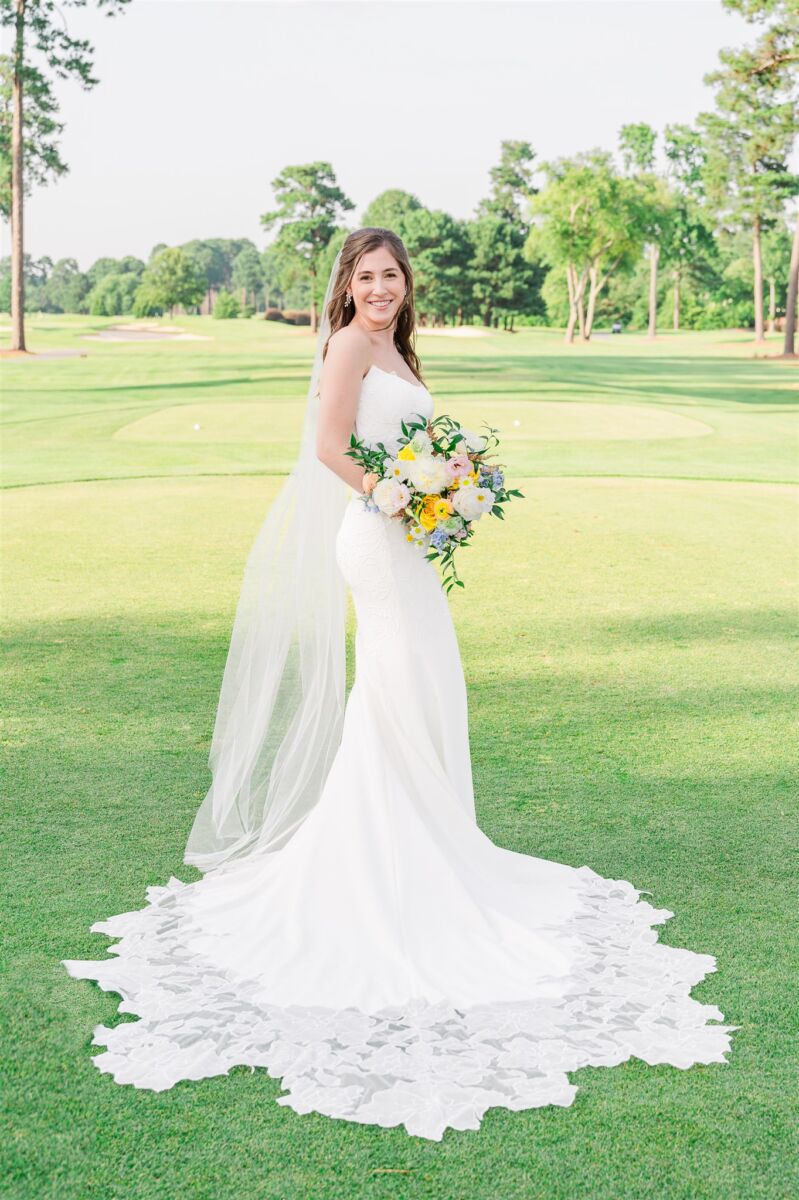 A bride smiles while standing on a golf course holding her large colorful bouquet thanks to her Wilmington wedding planners