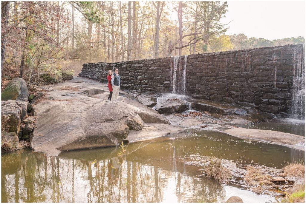A happy brunette couple standing under a waterfall during their Yates Mill engagement photo session by JoLynn Photography