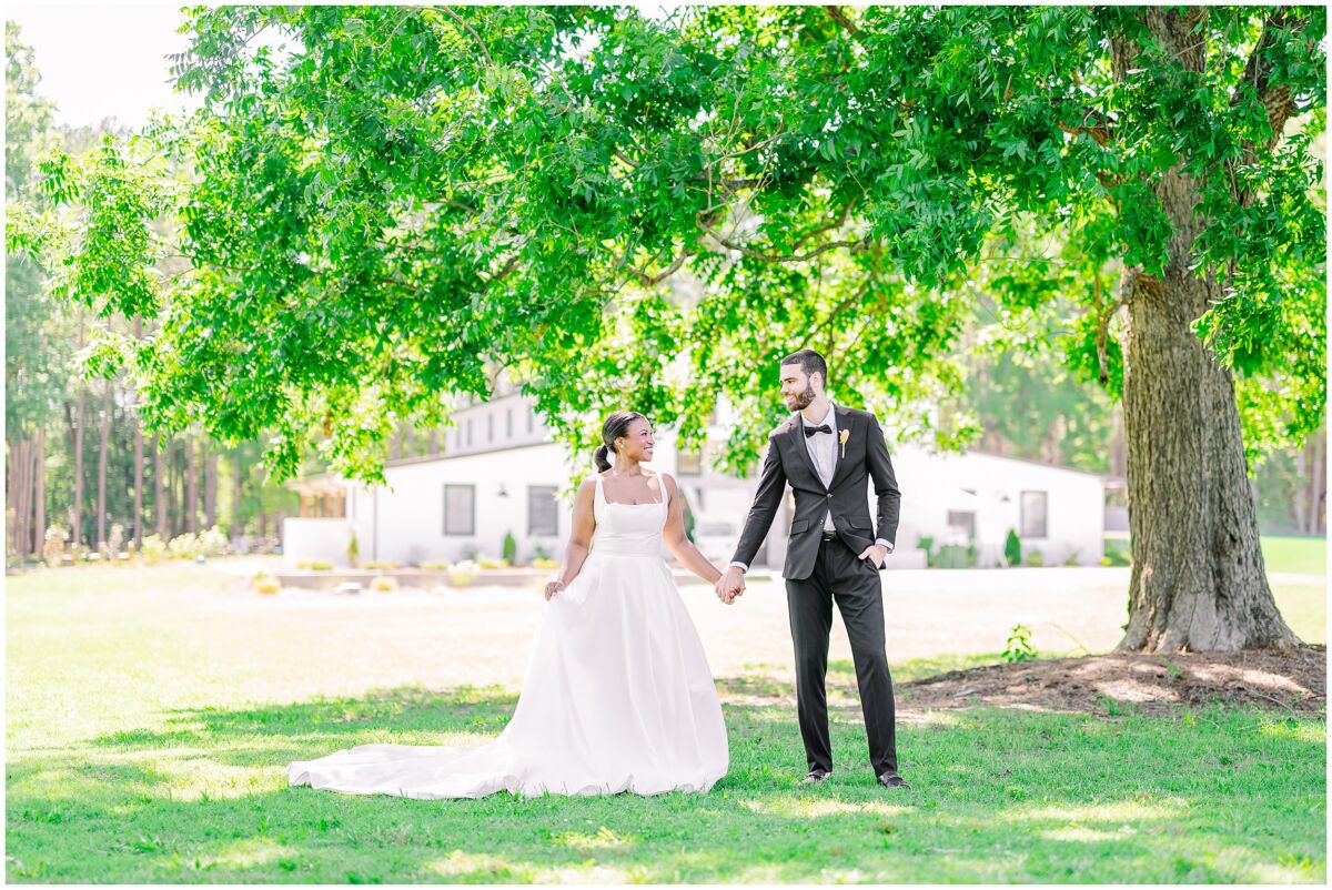 A beautiful couple holding hands under a vibrant green tree during their summer wedding at Stone Manor by JoLynn Photography